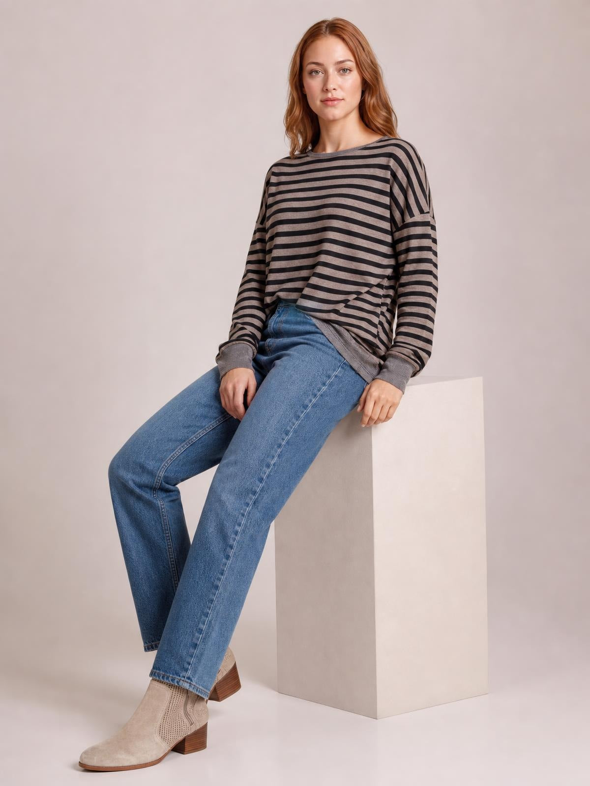 A model sits wearing a taupe and black striped round neck long sleeve T-shirt with a relaxed silhouette, ribbed cuffs, and side slit detail.