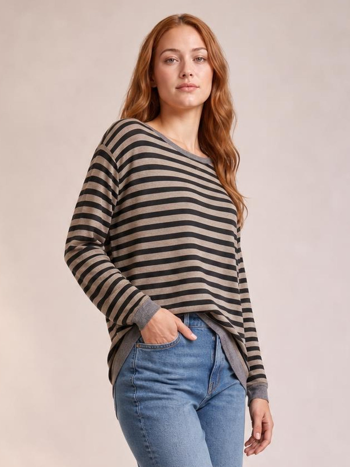 Model wearing a black and taupe striped round neck long sleeve T-shirt with a relaxed silhouette and visible side slit detail, posing with one hand in her jeans pocket.