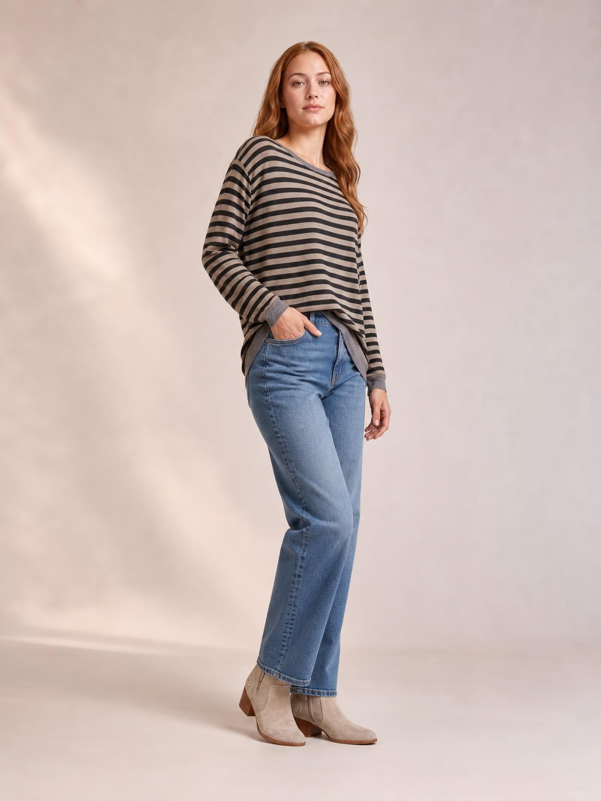 A woman poses in a charcoal and taupe striped round neck long sleeve T-shirt with a relaxed silhouette, side slit hem, and hands in her jeans pockets.
