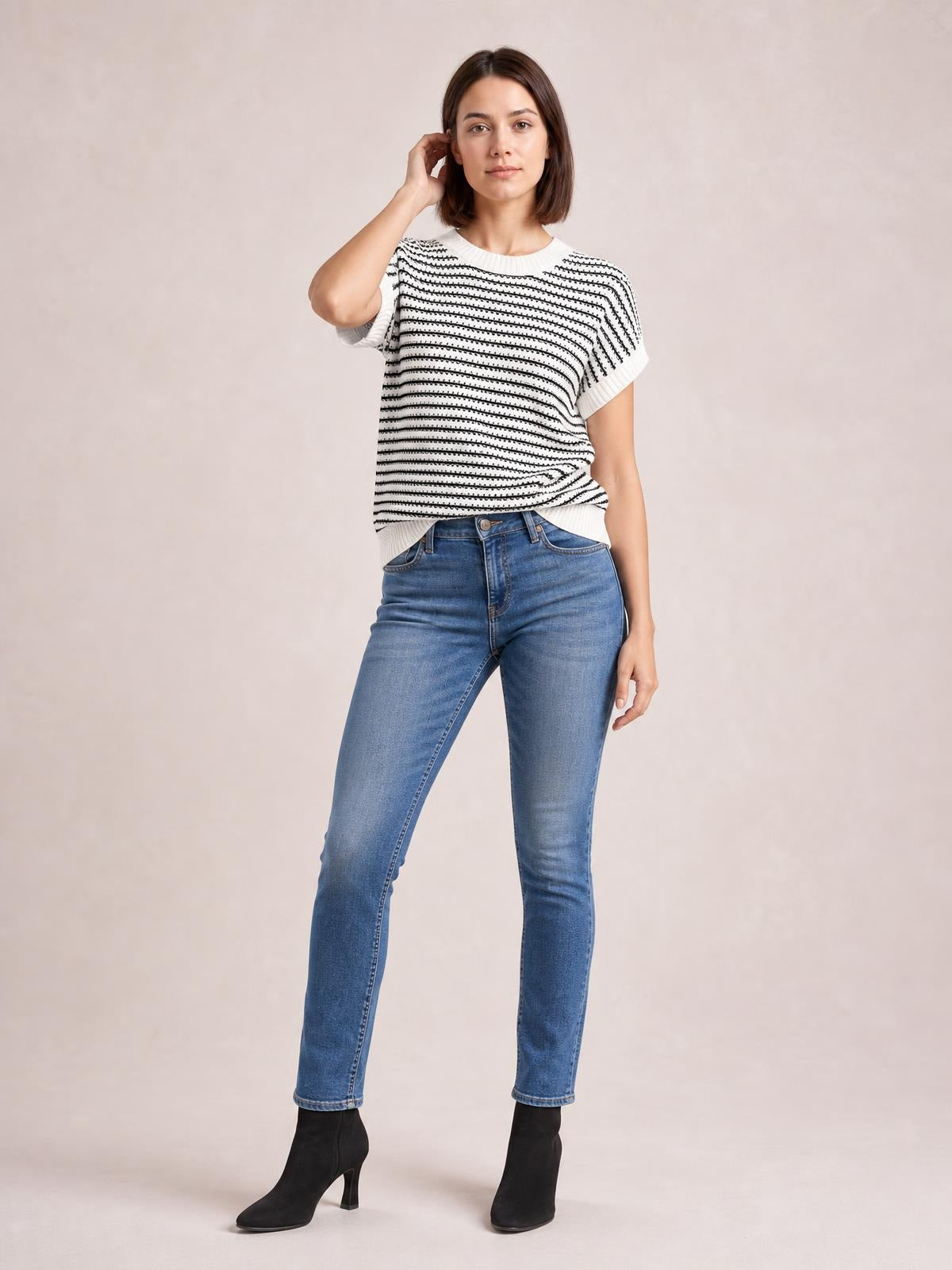 Model wearing a white and black striped round neck short sleeve knit top with ribbed trim and a relaxed silhouette, posing with one hand near her hair.