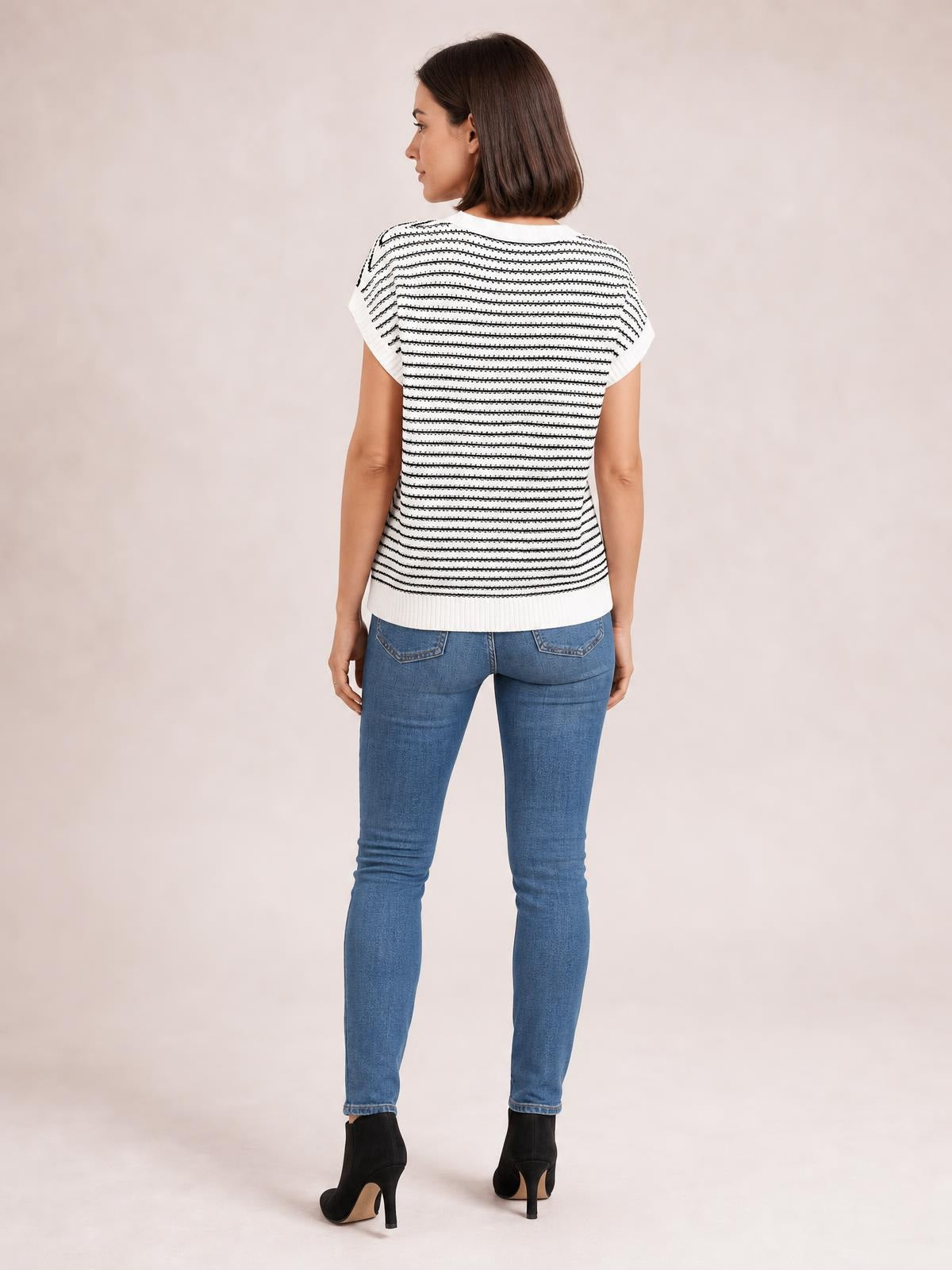 A woman stands facing away in a white and black striped knit top with a round neck, short sleeves, ribbed hem, and fitted silhouette.