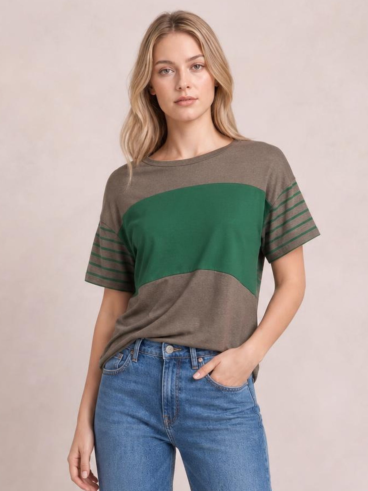 Model wears a loose striped round neck short sleeve T-shirt in gray and green with contrasting color-block panels, standing with one hand in her jeans pocket.