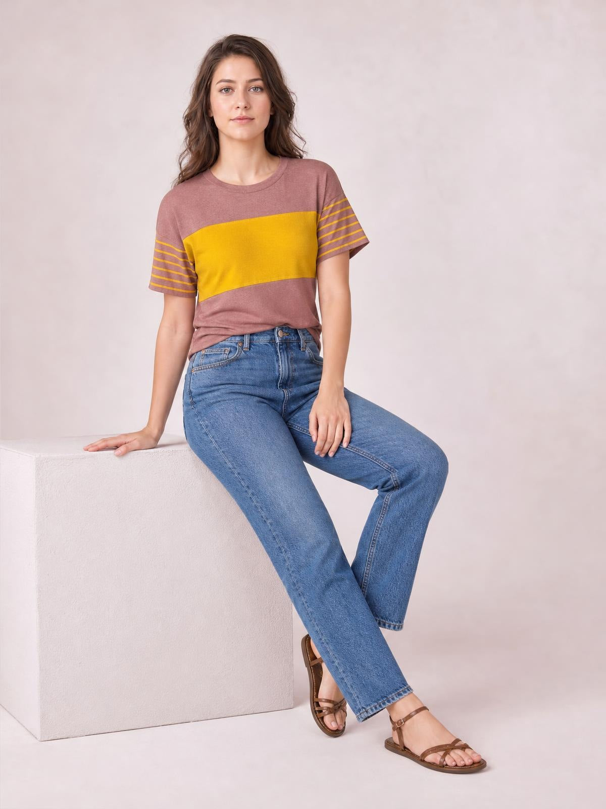 Model seated in a relaxed pose wears a mauve and mustard striped round neck short sleeve T-shirt with a straight silhouette and contrast sleeve stripes.