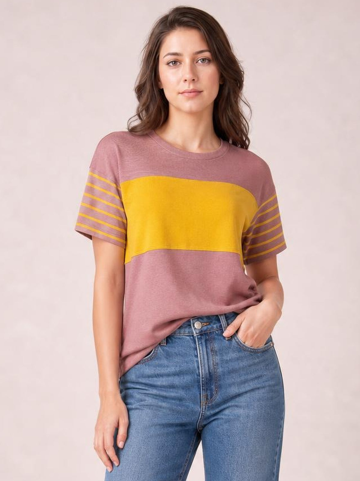 Model wearing a mauve and mustard striped round neck short sleeve T-shirt with a color-blocked front and relaxed silhouette, posing with one hand in her jeans pocket.
