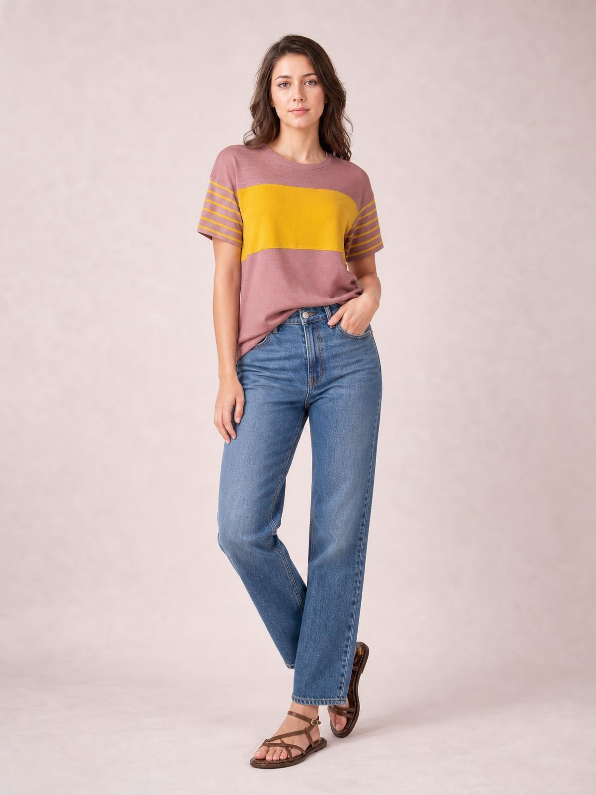Model wears a mauve and mustard striped round neck short sleeve T-shirt with a relaxed silhouette, posing with one hand in her jeans pocket.