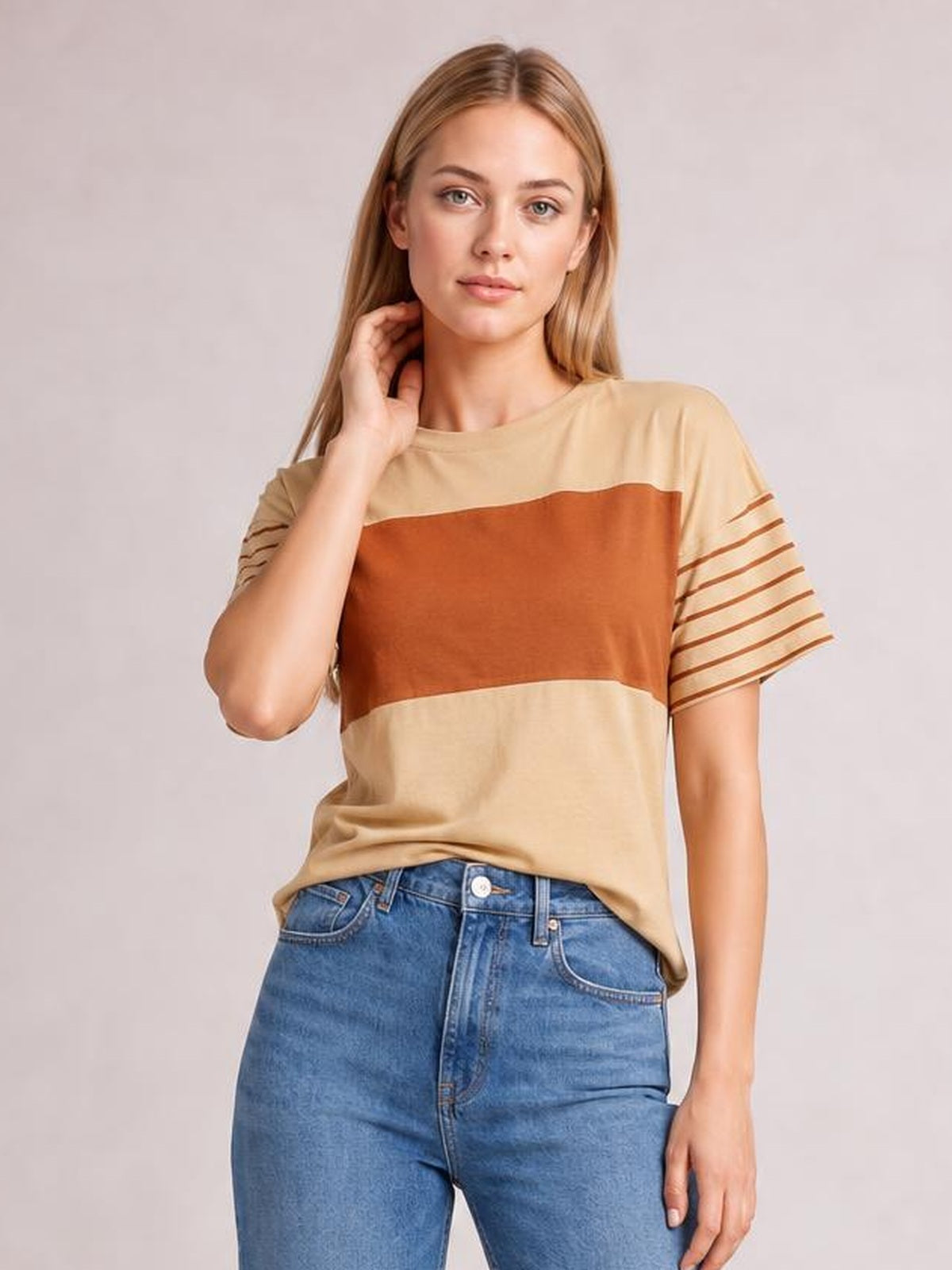Model poses with one hand near her neck, wearing a beige round neck short sleeve T-shirt with a rust color block and striped sleeves in a relaxed silhouette.