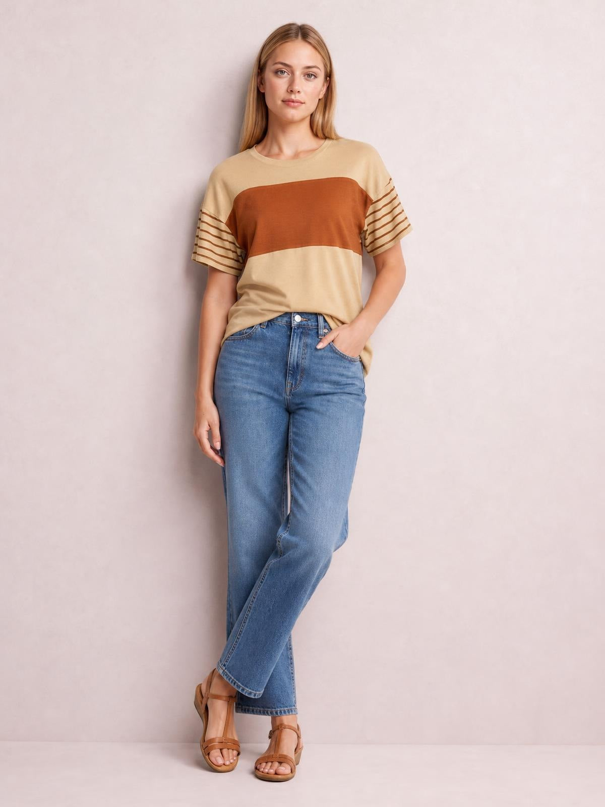 A woman poses in a beige and rust striped round neck short sleeve T-shirt with a relaxed silhouette, paired with jeans and one hand in a pocket.