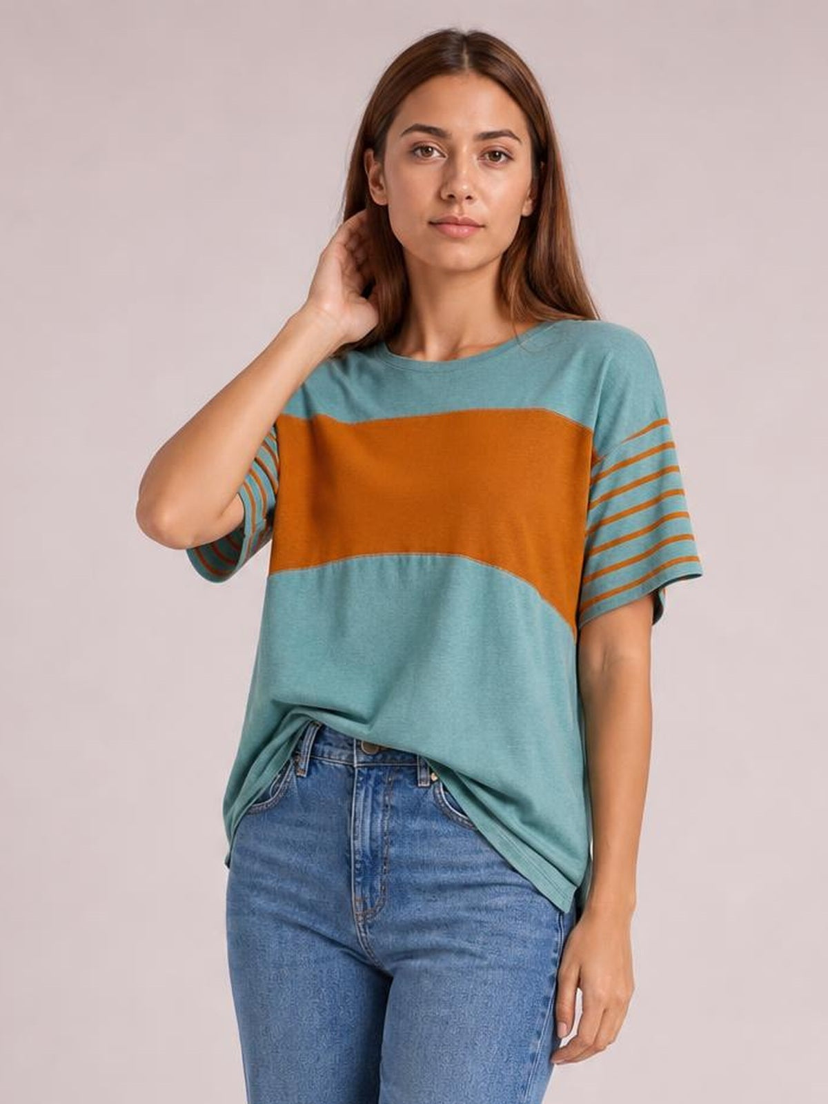 Model wearing a loose-fit teal striped round neck short sleeve T-shirt with a wide rust-orange chest stripe, standing with one hand touching her hair.
