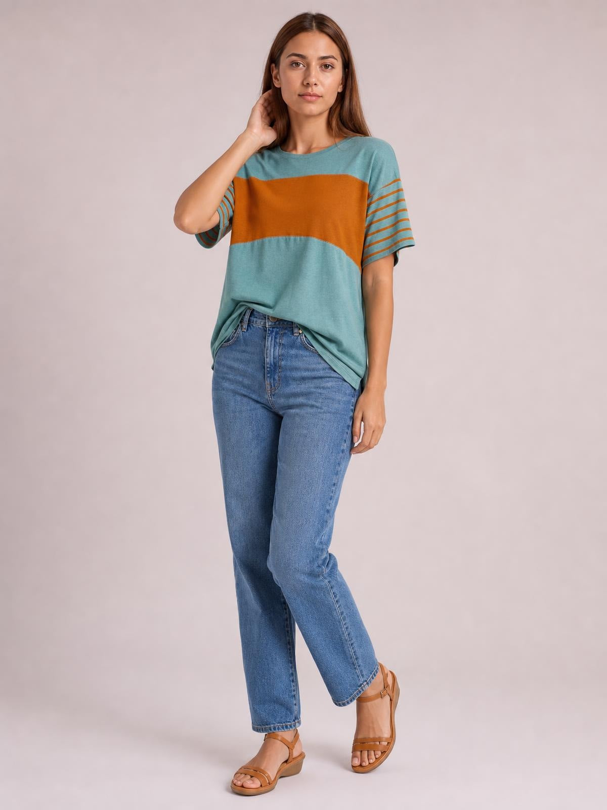 Model wearing a teal striped round neck short sleeve T-shirt with a relaxed silhouette, orange color-block band, and striped sleeves, posing with one hand near her hair.