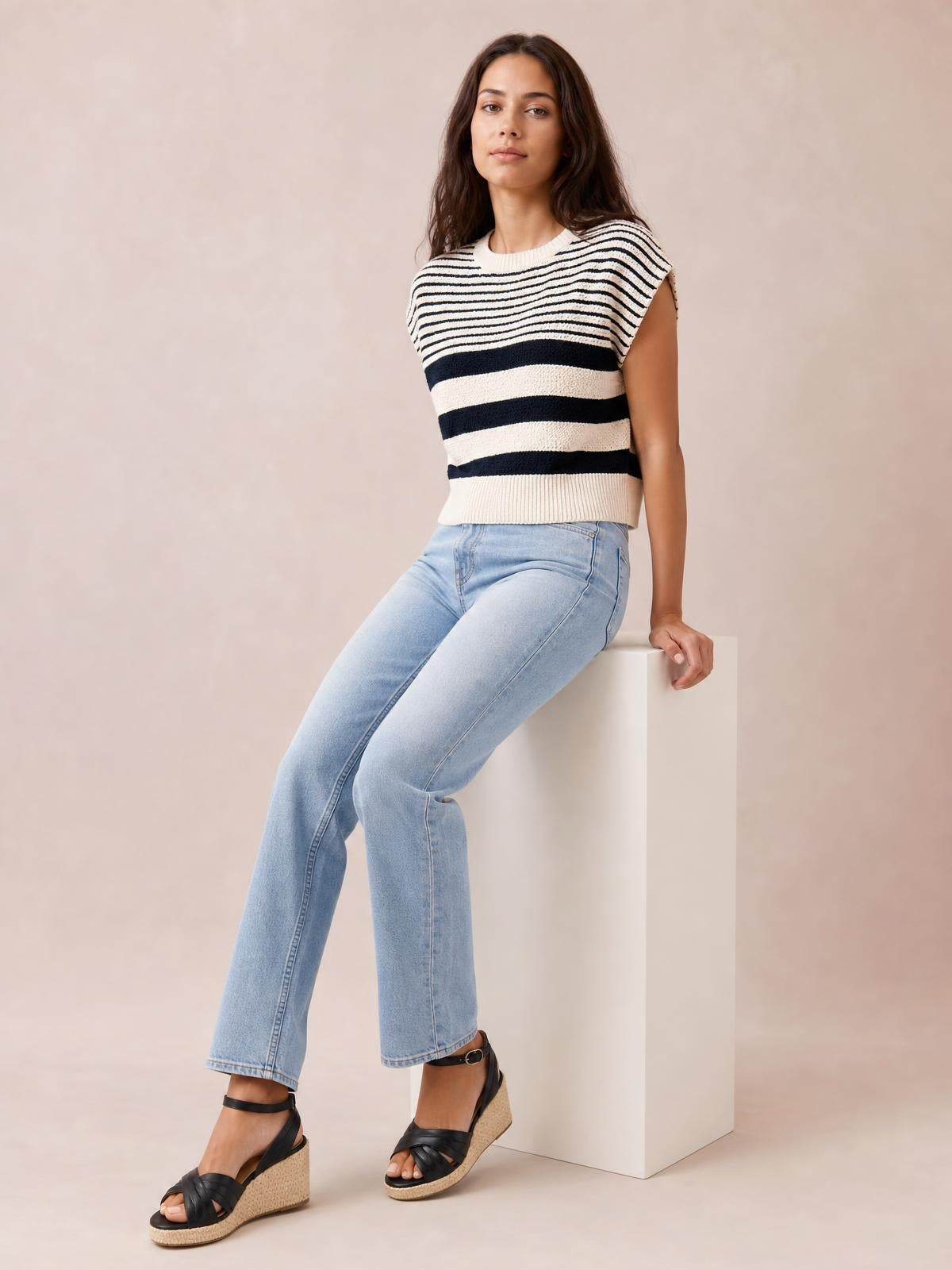 Model seated in a relaxed pose wears a black and cream striped round neck sweater vest with a sleeveless silhouette, ribbed neckline, hem, and side seams.