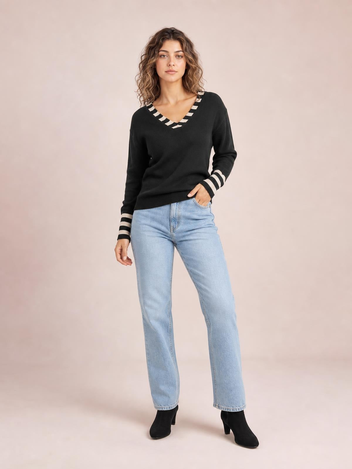 A model stands with one hand in her jeans pocket, wearing a black drop-shoulder sweater with white striped V-neck trim, cuffs, and a ribbed hem.