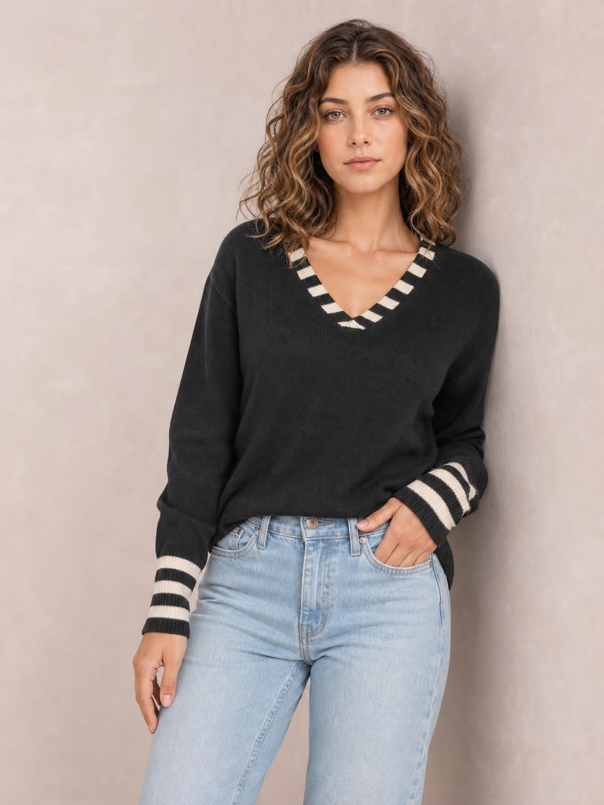 Model poses in a black drop shoulder sweater with a V-neck and striped trim at the neckline and cuffs, paired with light-wash jeans.