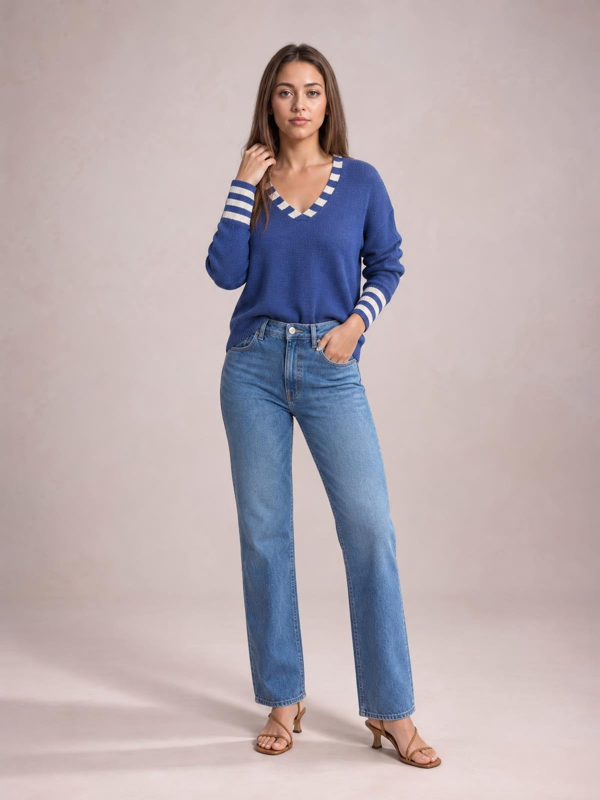 Model poses in a blue drop shoulder sweater with white striped V-neck and cuff trim, relaxed silhouette, long sleeves, and jeans.