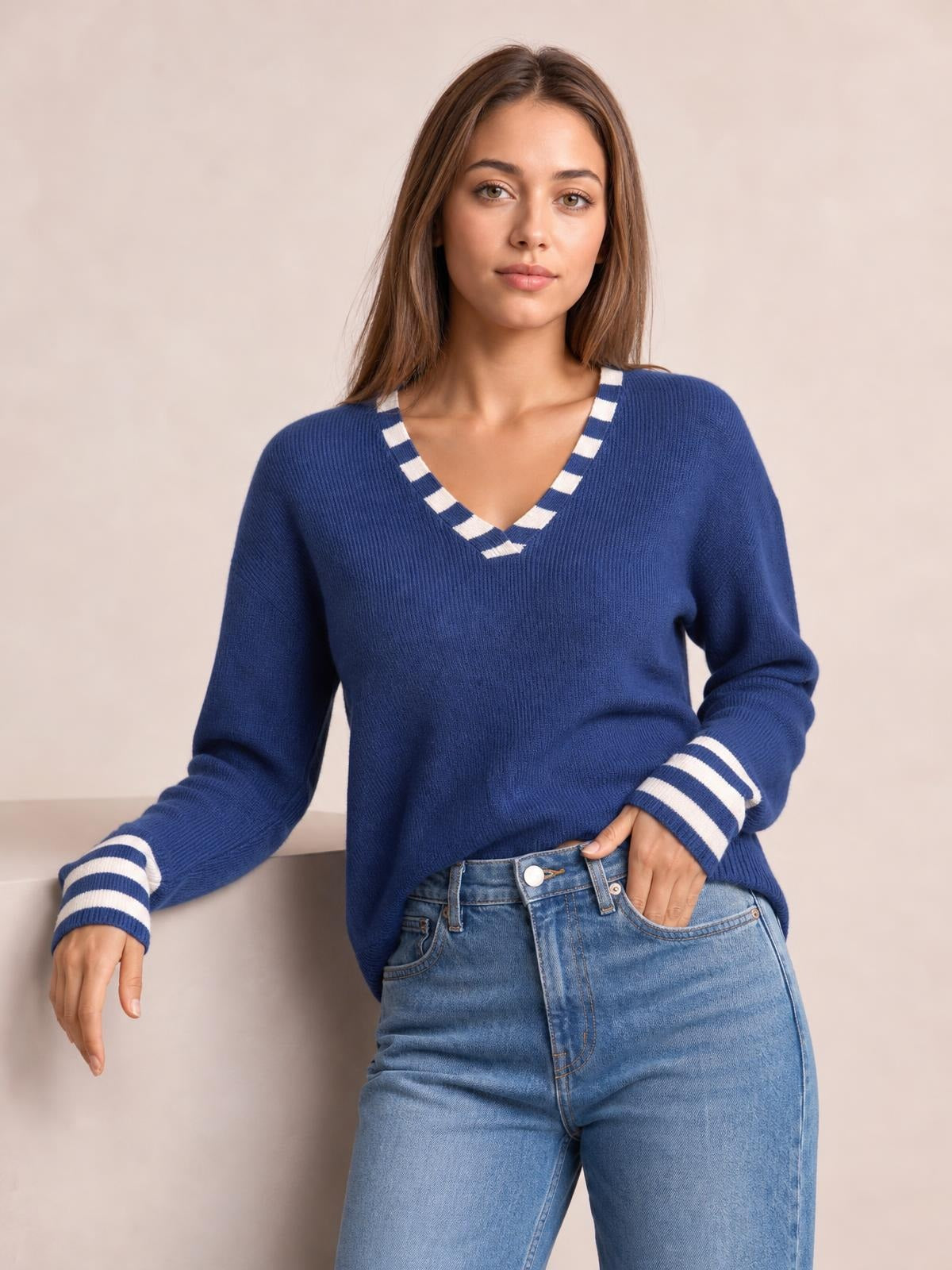 Model wears a navy blue drop-shoulder sweater with white striped V-neck trim and cuffs, a relaxed silhouette, and one hand resting in her jeans pocket.