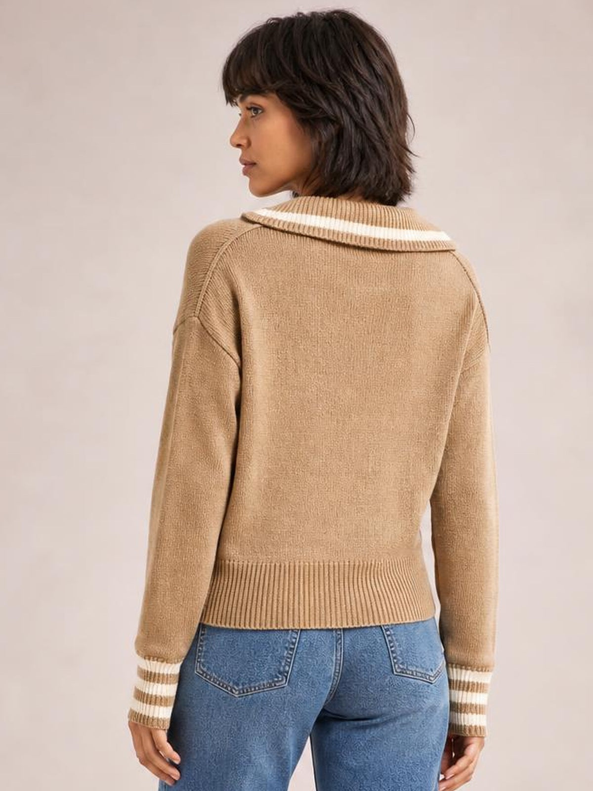 A model looks over her shoulder wearing a relaxed tan drop-shoulder sweater with striped trim at the collar and cuffs and a ribbed hem.
