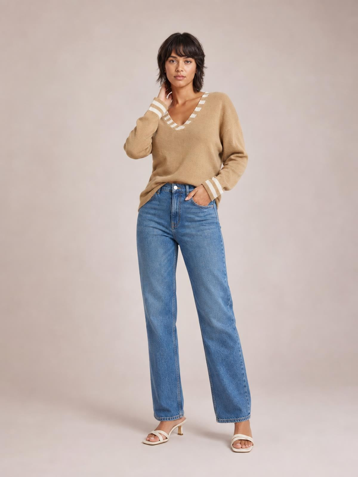 Model poses with one hand at her neck wearing a tan drop-shoulder sweater with striped V-neck and cuff trim and a relaxed silhouette.