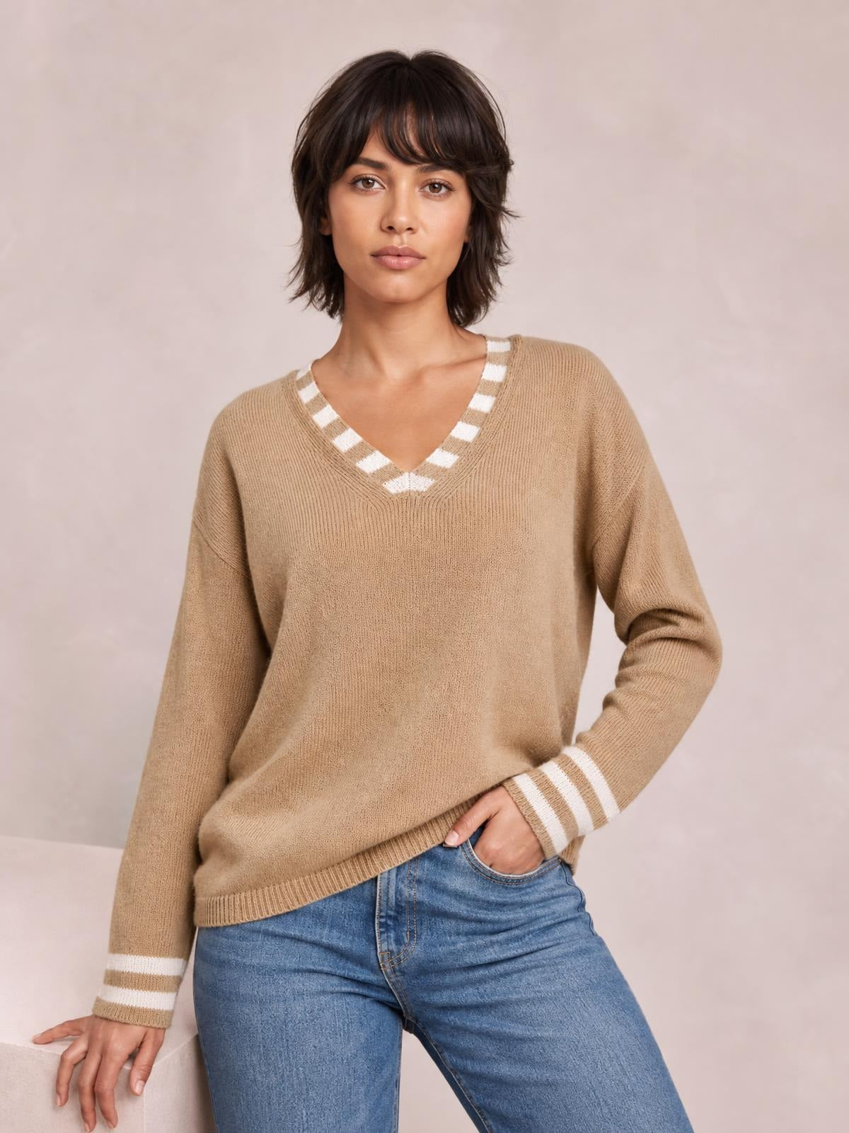 Model wearing a camel drop-shoulder sweater with a relaxed silhouette, V-neckline, and contrasting striped trim at the collar and cuffs.