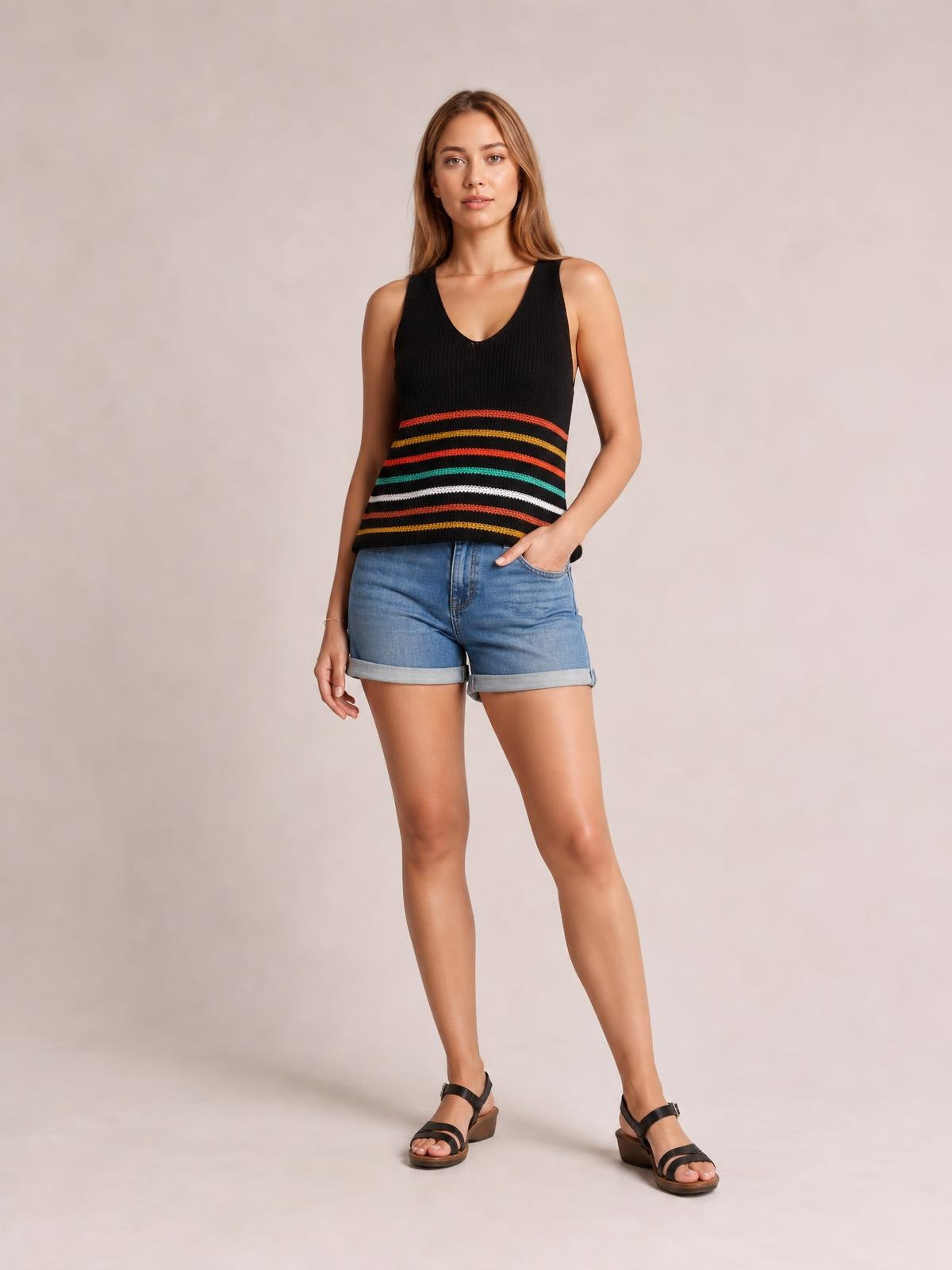 Model wearing a black striped V-neck knit tank with a sleeveless, relaxed silhouette and multicolored horizontal bands, posed with one hand in a pocket.