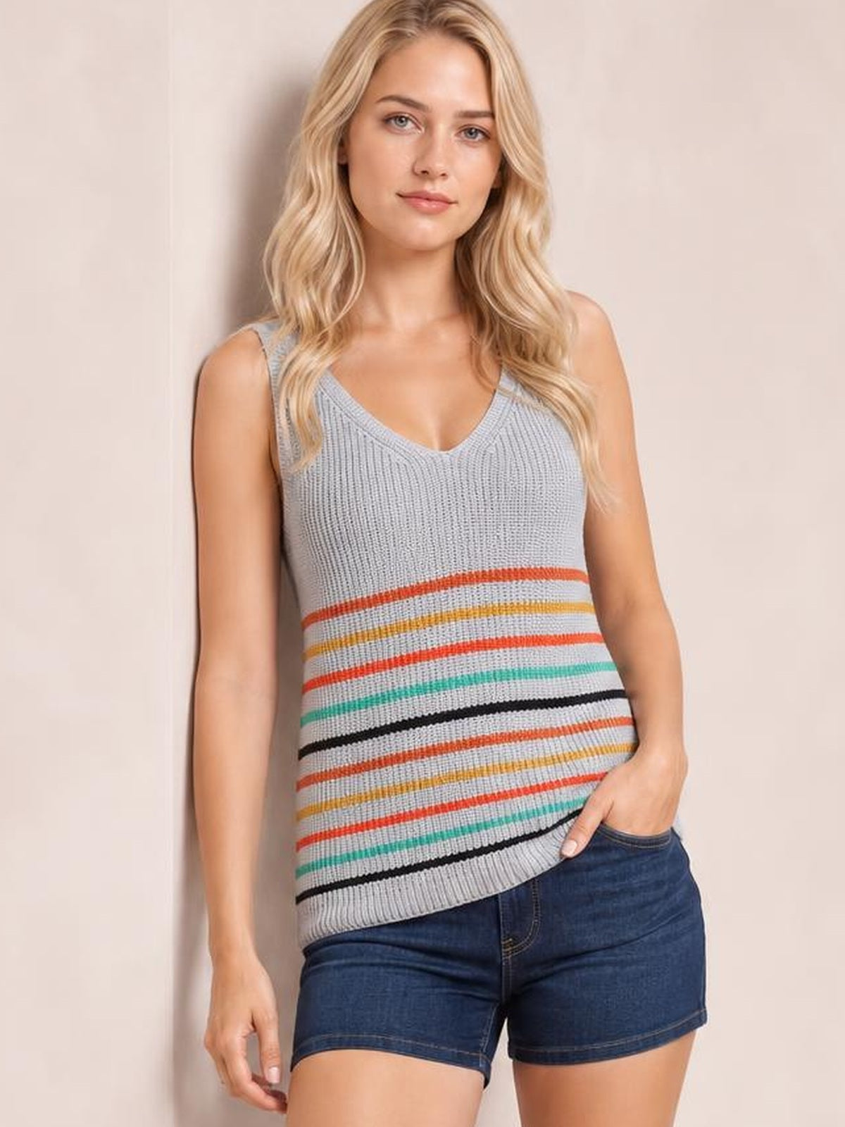 Model wears a gray striped V-neck knit tank with colorful horizontal bands, a fitted straight silhouette, and wide shoulder straps, posing with one hand in her pocket.