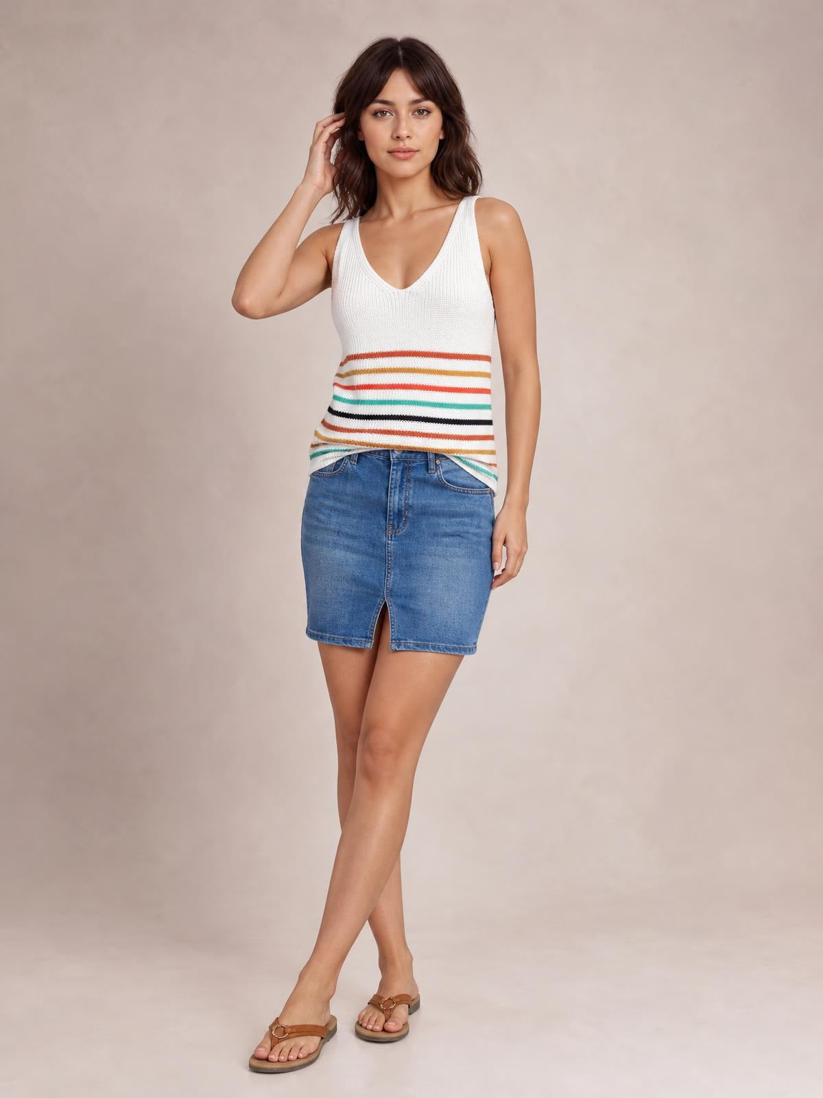 Model poses in a white striped V-neck knit tank with multicolor horizontal bands, sleeveless silhouette, ribbed texture, and a straight hem over a denim skirt.