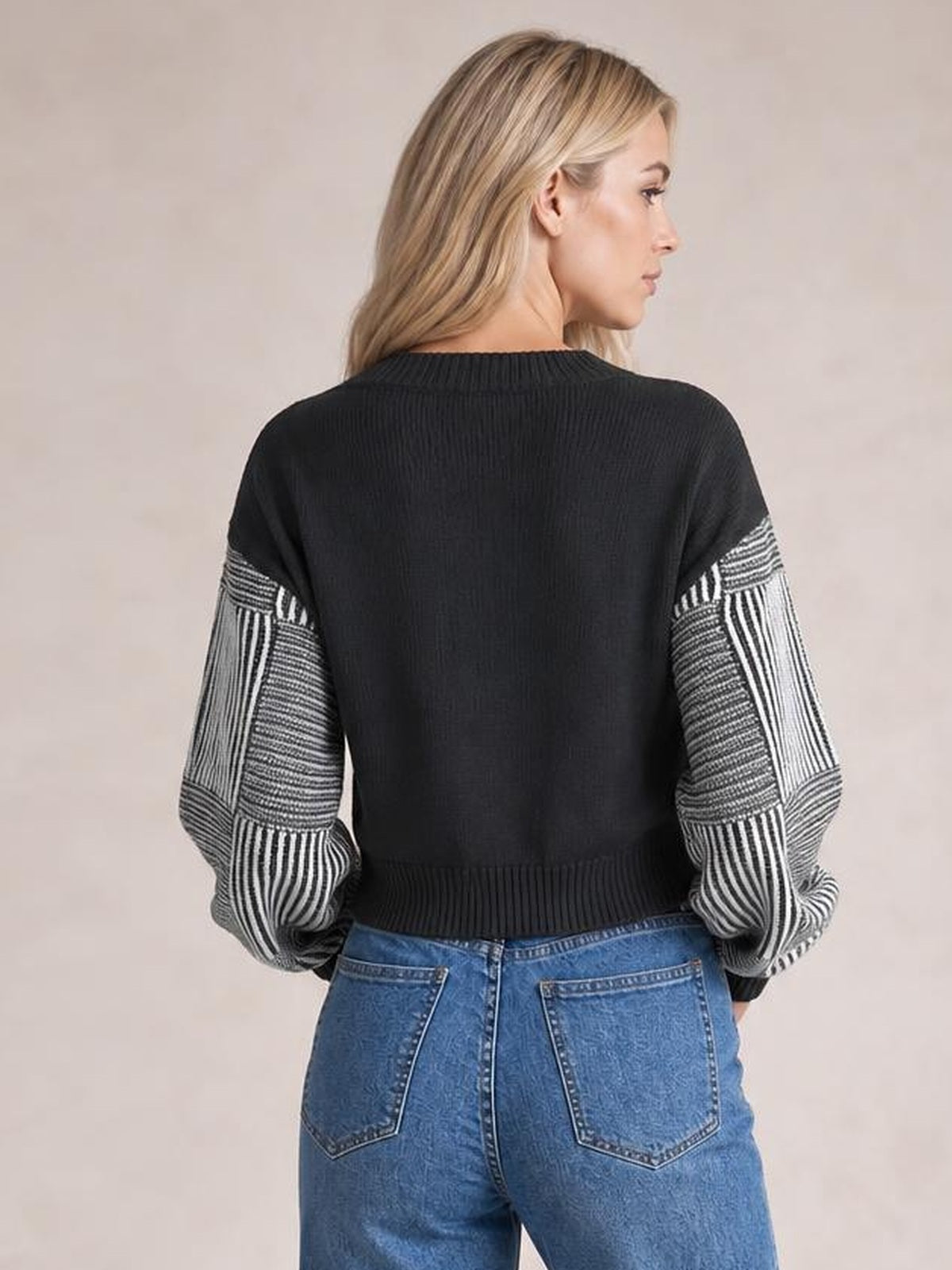 A model stands from behind wearing a black long-sleeve sweater with contrasting striped sleeves, a ribbed crew-style neckline, cuffs, and hem.