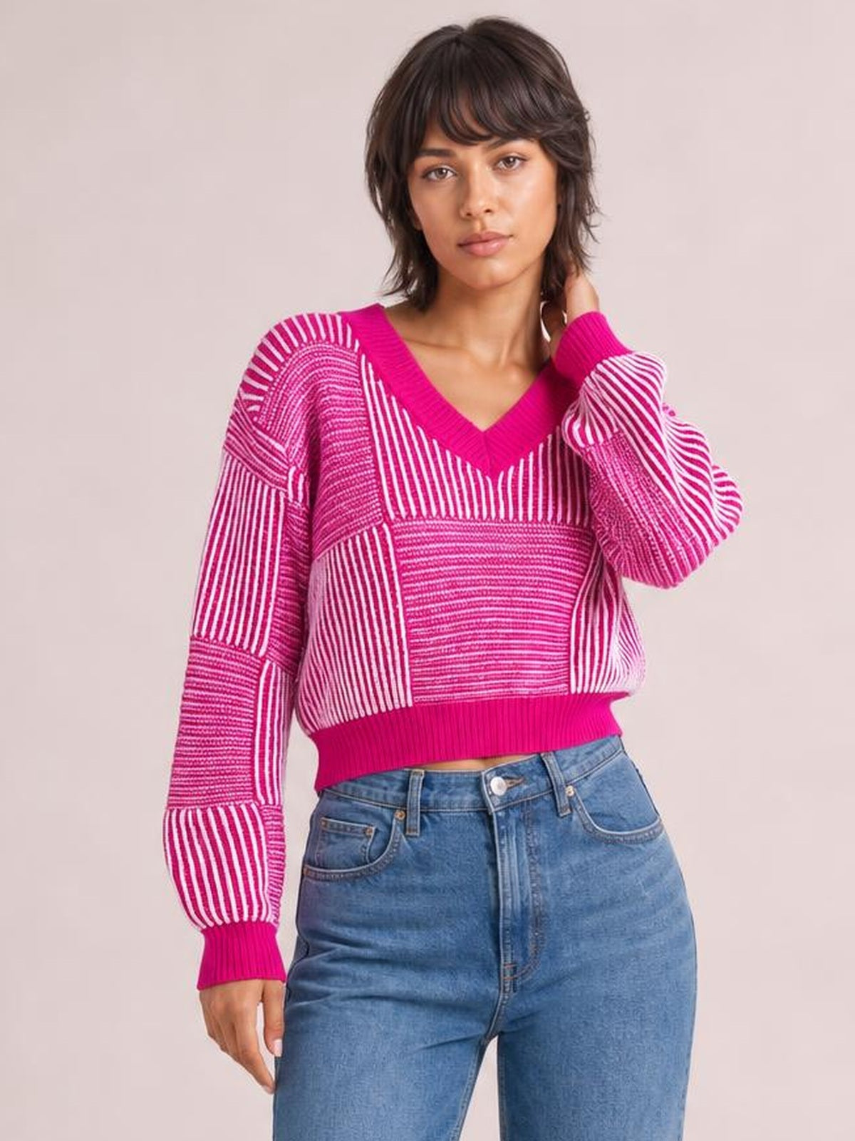 A model poses in a pink and white striped V-neck long-sleeve sweater with ribbed trim, a cropped silhouette, and contrasting directional knit stripes.
