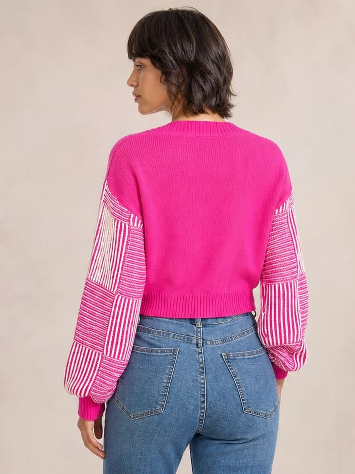 Model shown from behind wearing a bright pink long sleeve sweater with contrasting striped sleeves, ribbed collar, cuffs, hem, and a cropped silhouette.