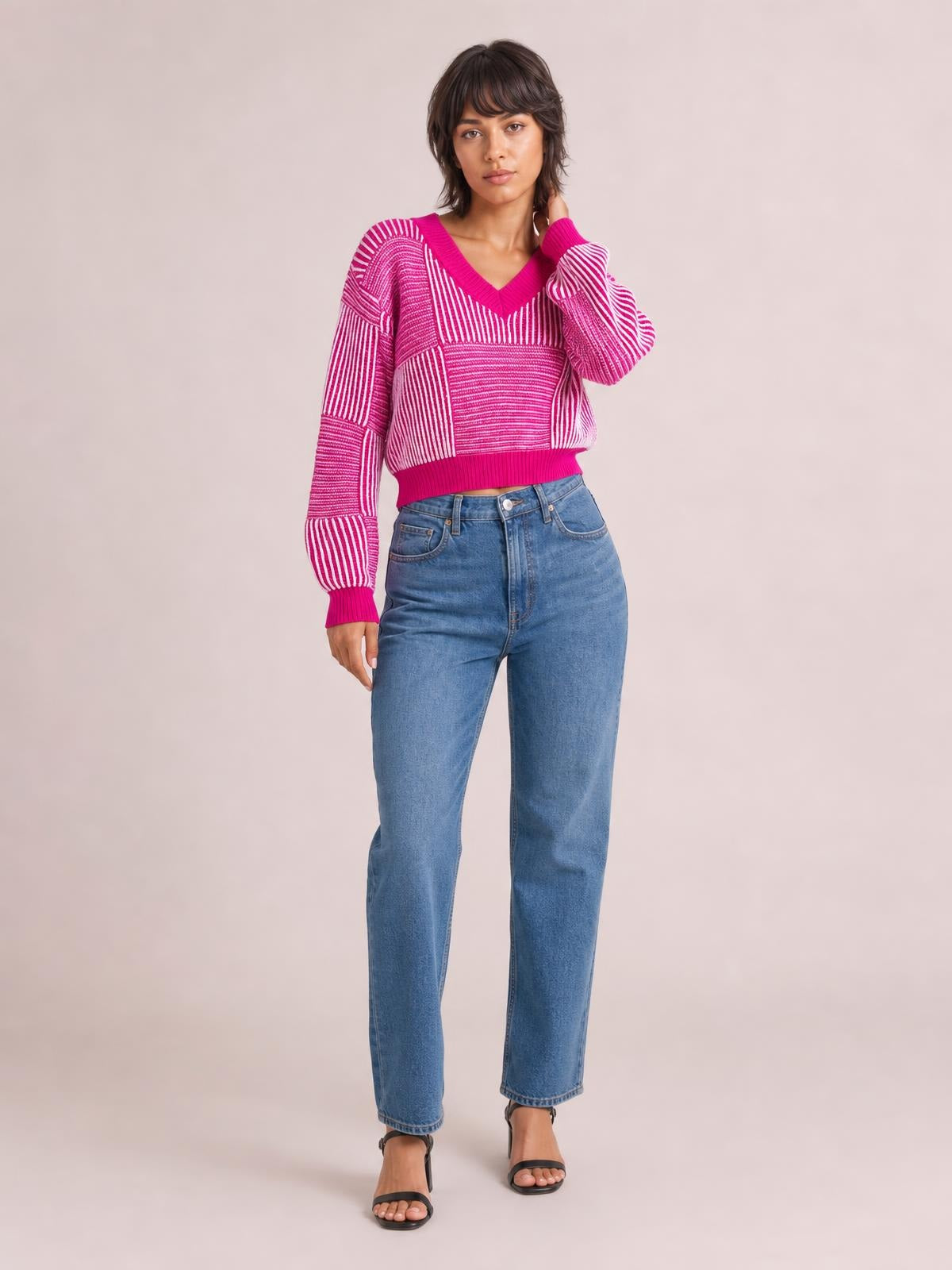 Model wearing a pink and white striped V-neck long sleeve sweater with a relaxed cropped silhouette, ribbed trim, and one hand resting near her neck.