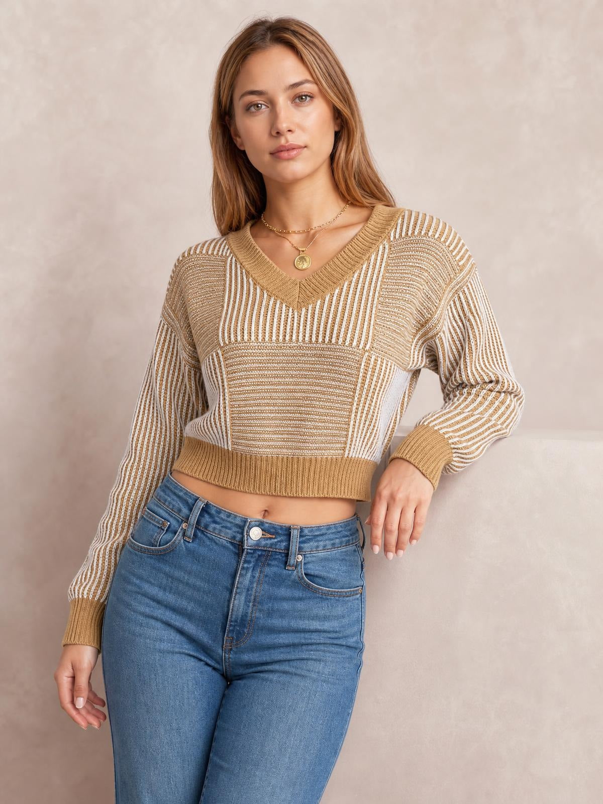 Model wearing a tan and white striped V-neck long sleeve sweater with a cropped silhouette, ribbed neckline, cuffs, and hem, posing with one hand on her hip.