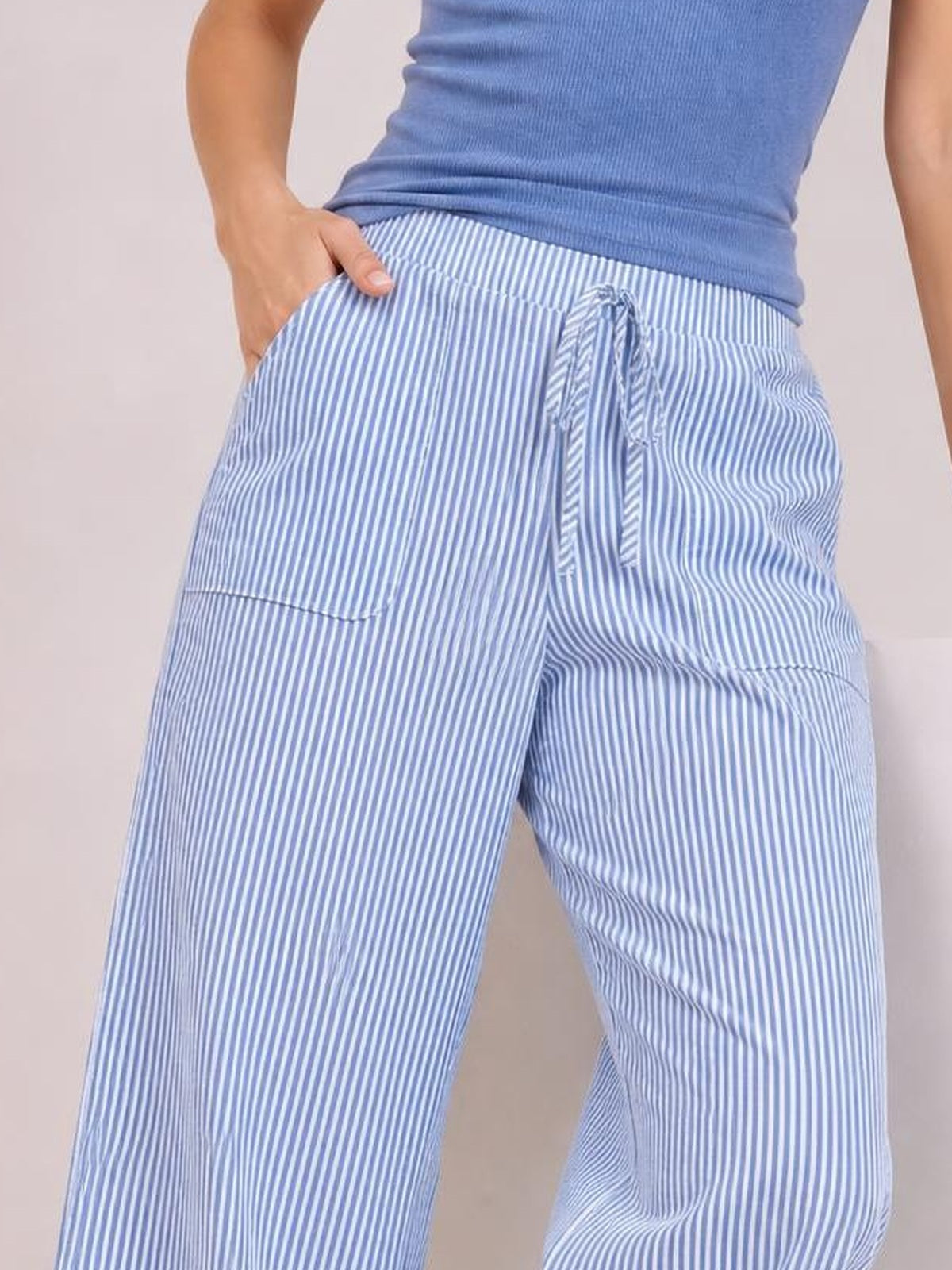 Model wearing light blue and white striped wide leg pants with a drawstring waist, side pockets, and relaxed flowing legs, posing with one hand in a pocket.