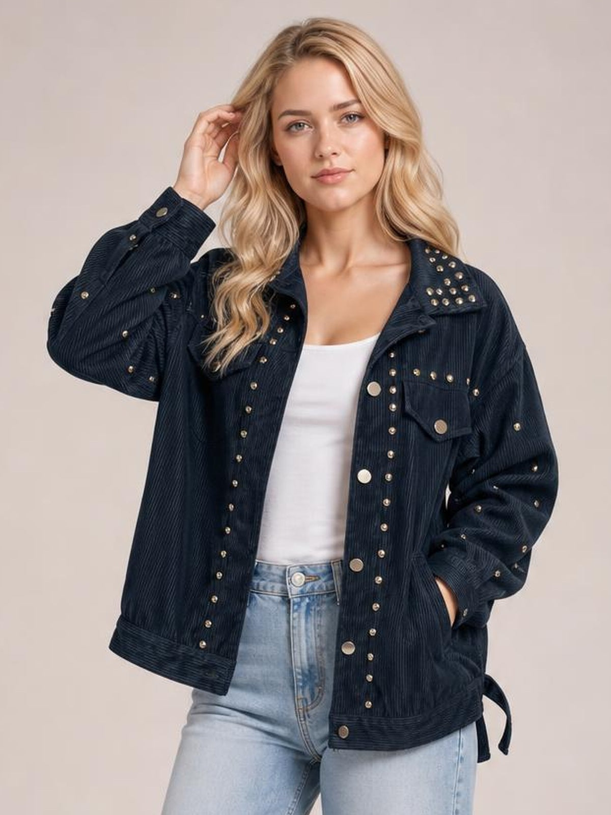 Model wears a relaxed dark navy collared button-up jacket with studded detailing, flap pockets, long sleeves, and a textured finish while posing with one hand in a pocket.
