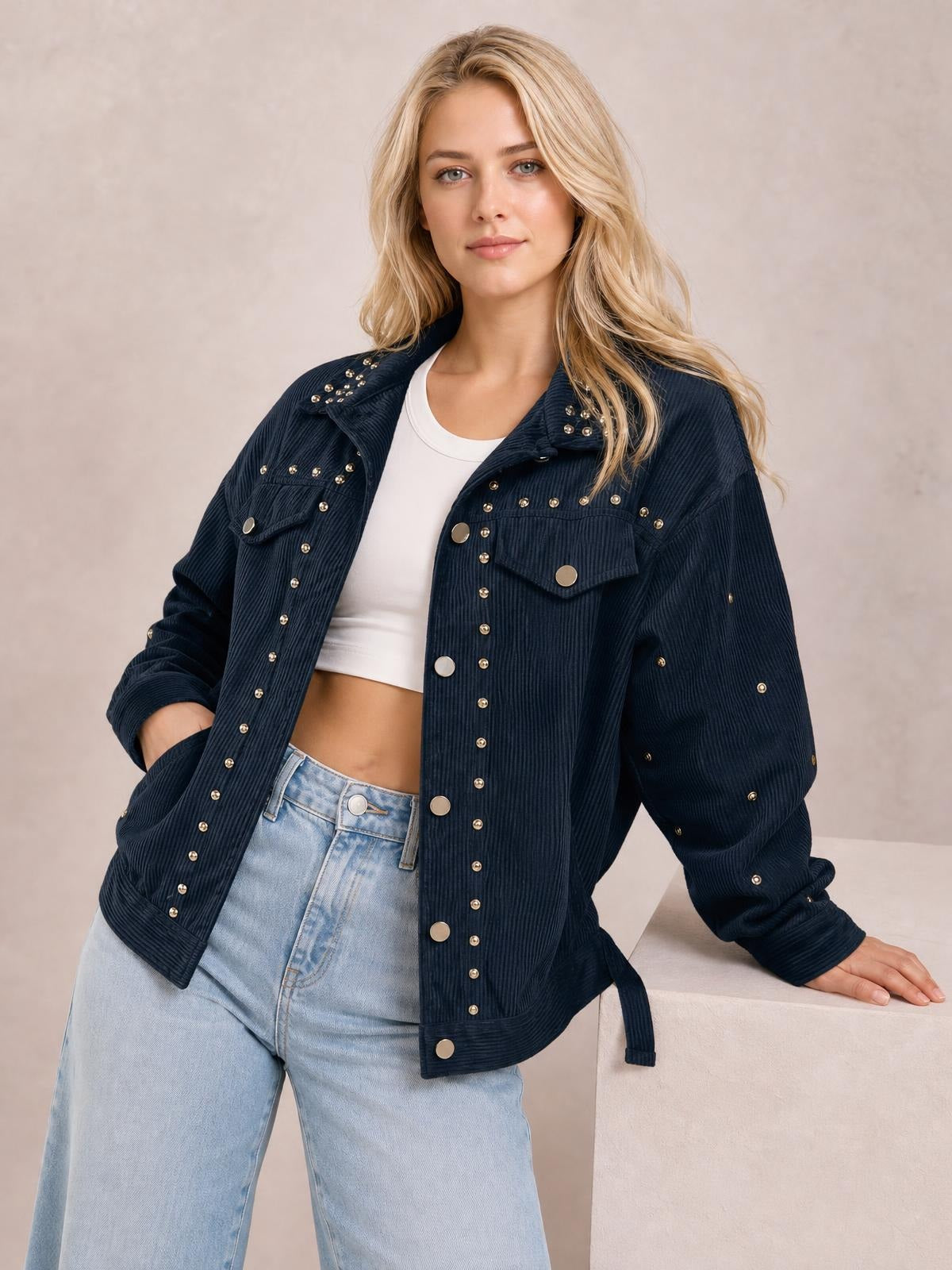 Model wears a navy studded collared button-up jacket with a relaxed silhouette, patch pockets, ribbed texture, and long sleeves, posing with one hand in a pocket.