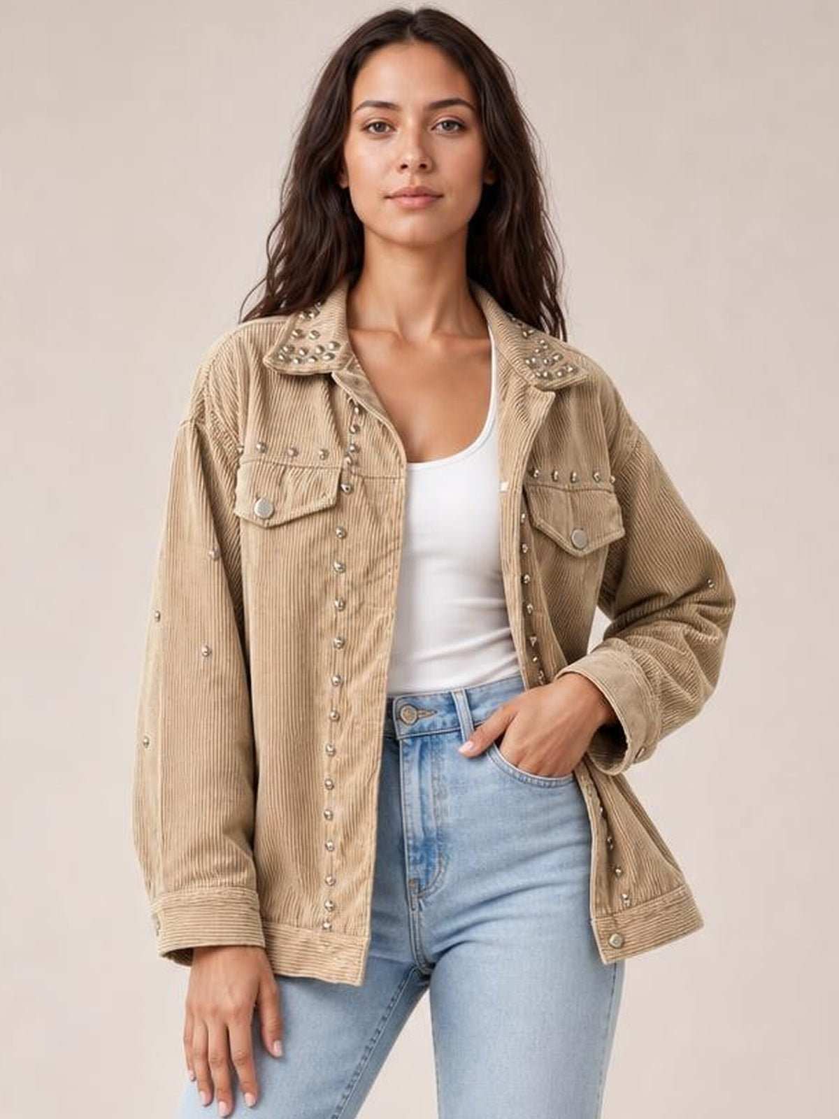 A model poses in a relaxed tan studded collared button-up jacket with chest pockets, long sleeves, and a straight silhouette over a white top.