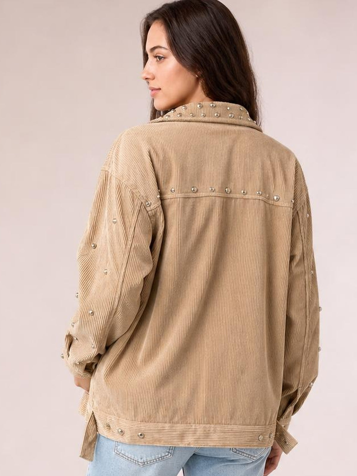 A woman looks over her shoulder wearing a tan studded collared button-up jacket with a relaxed silhouette, ribbed texture, and studded seams, cuffs, and hem.