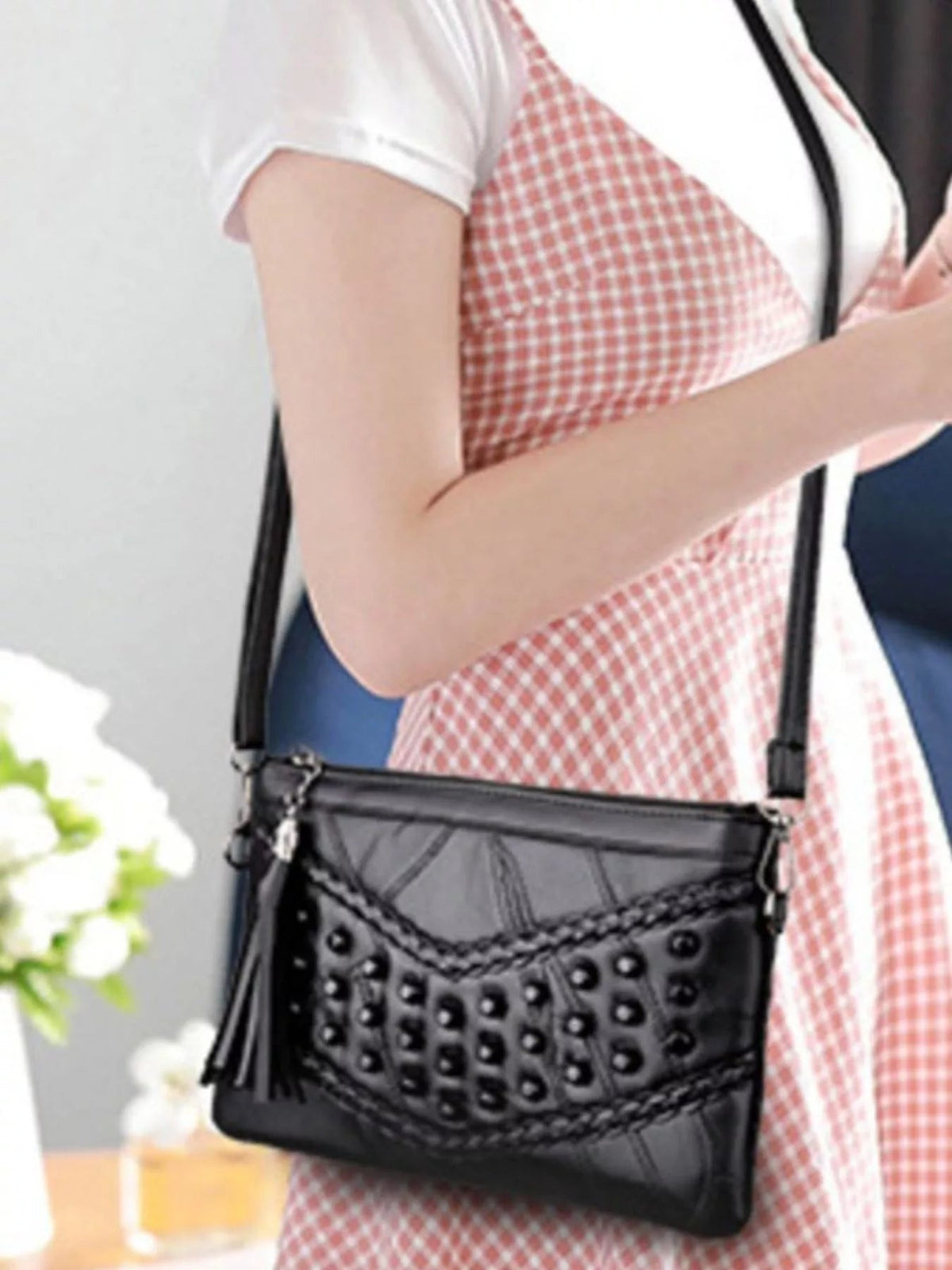 A model wears a medium black PU leather crossbody bag with a rectangular silhouette, studded front, braided trim, and adjustable strap while holding her arms across it.