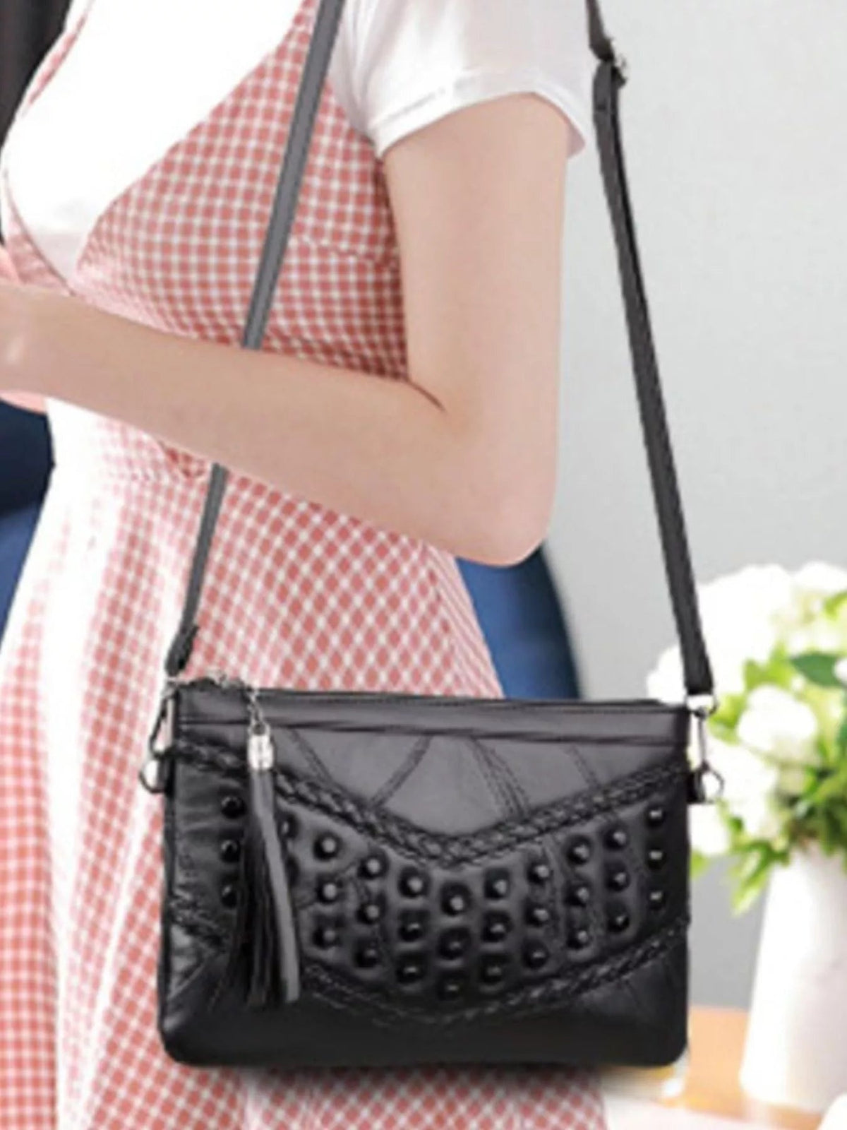 A model poses with a medium black PU leather crossbody bag featuring a rectangular silhouette, studded front panel, braided trim, and long shoulder strap.