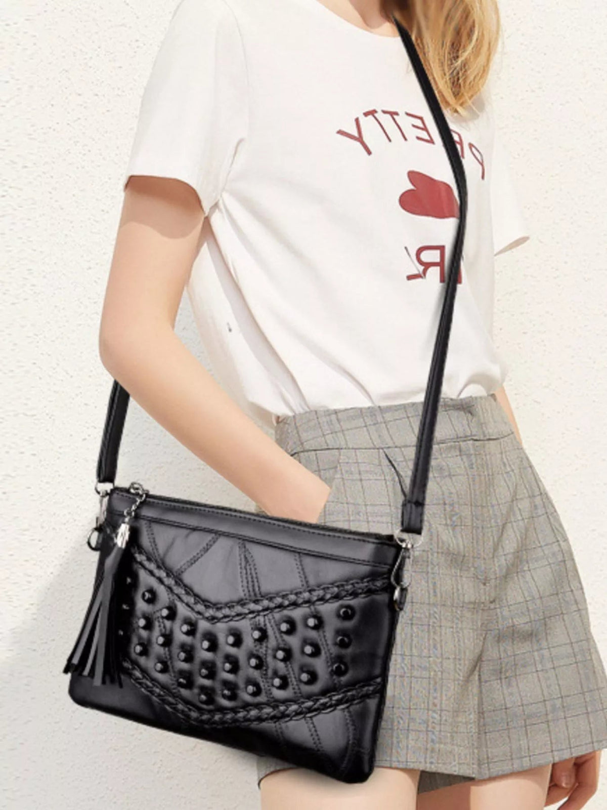 A model wears a medium black PU leather-look crossbody bag with studded panels, braided trim, tassel detail, and a long adjustable strap.