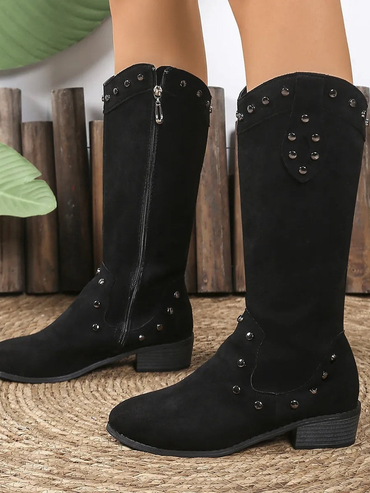 Model wearing black studded mid-calf boots with low block heels, rounded toes, textured panels, and visible side zipper closures.