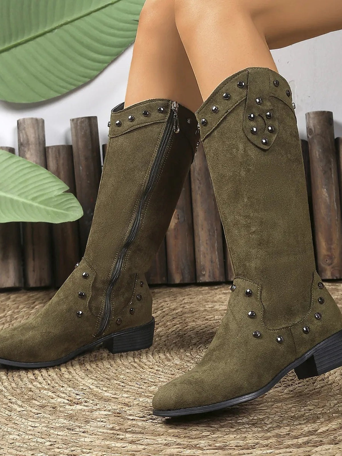 A model poses in olive green studded mid-calf boots with decorative paneling, side zipper closures, pointed toes, and low block heels.