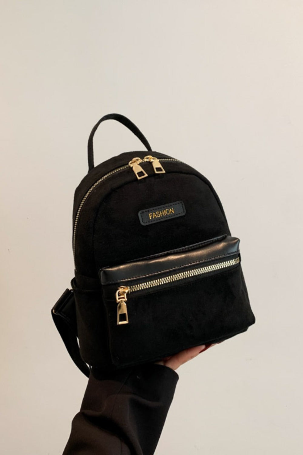 A model holds a small black suede backpack with a rounded silhouette, top handle, adjustable straps, front zip pocket, and gold-tone zippers.