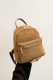 A model holds a small tan suede backpack with a softly structured silhouette, top handle, adjustable straps, and multiple front zippered compartments.