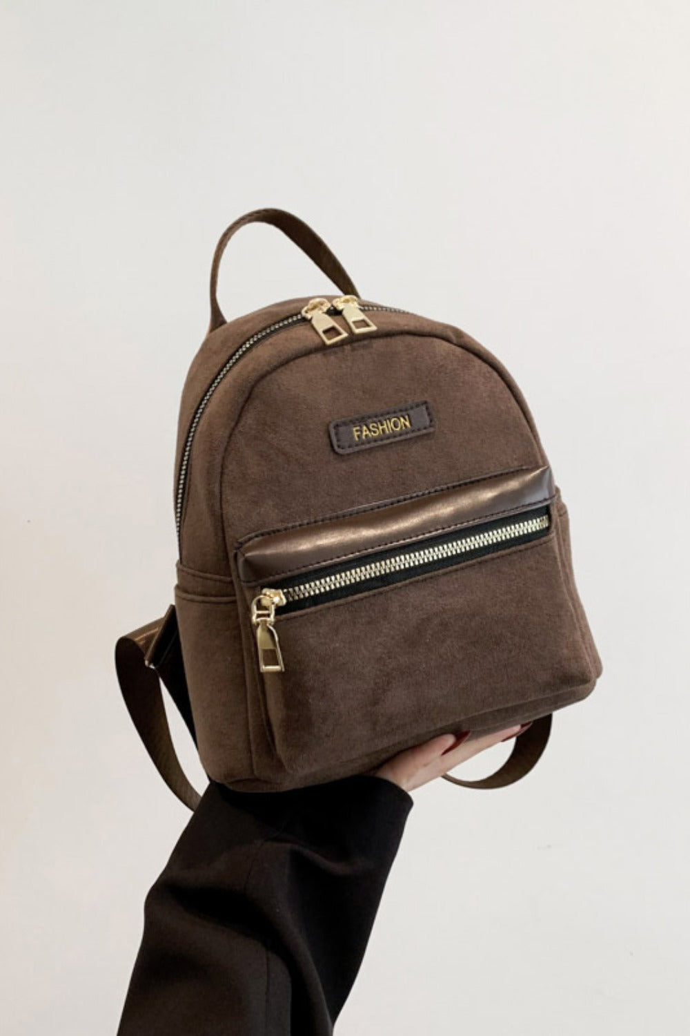 A model holds a small brown suede backpack with a softly structured silhouette, top handle, adjustable straps, gold-tone zippers, and a front zip pocket.