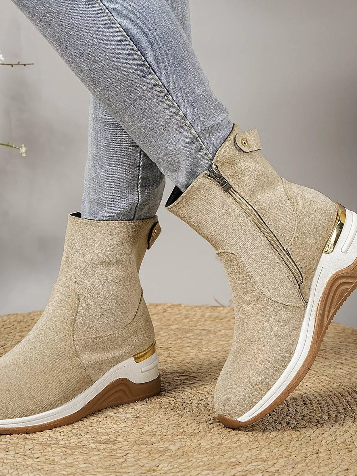 Beige suede ankle boots feature side zippers, buckle straps, mid-height wedge heels, and contrasting white rubber soles, shown on crossed legs.