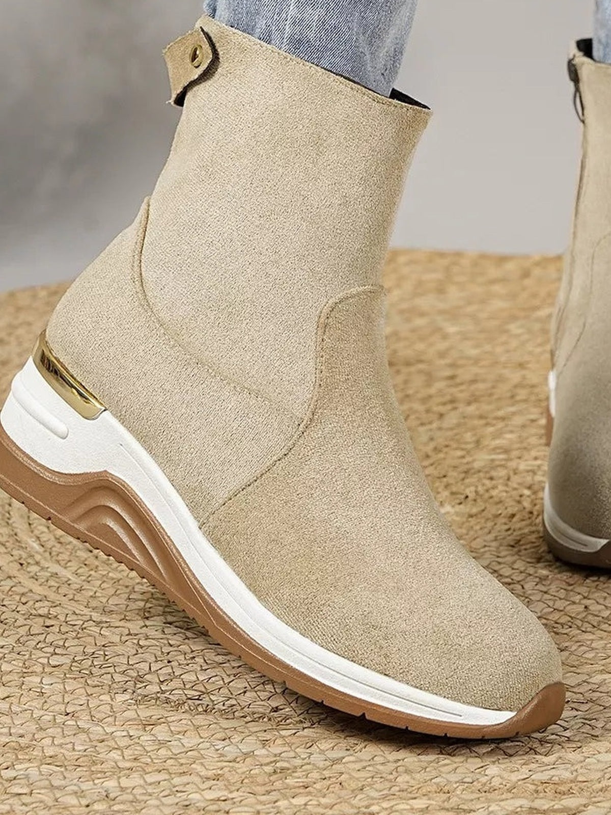A model wears a beige suede ankle boot with a rounded toe, mid-height wedge heel, white midsole, and tan rubber outsole in side view.