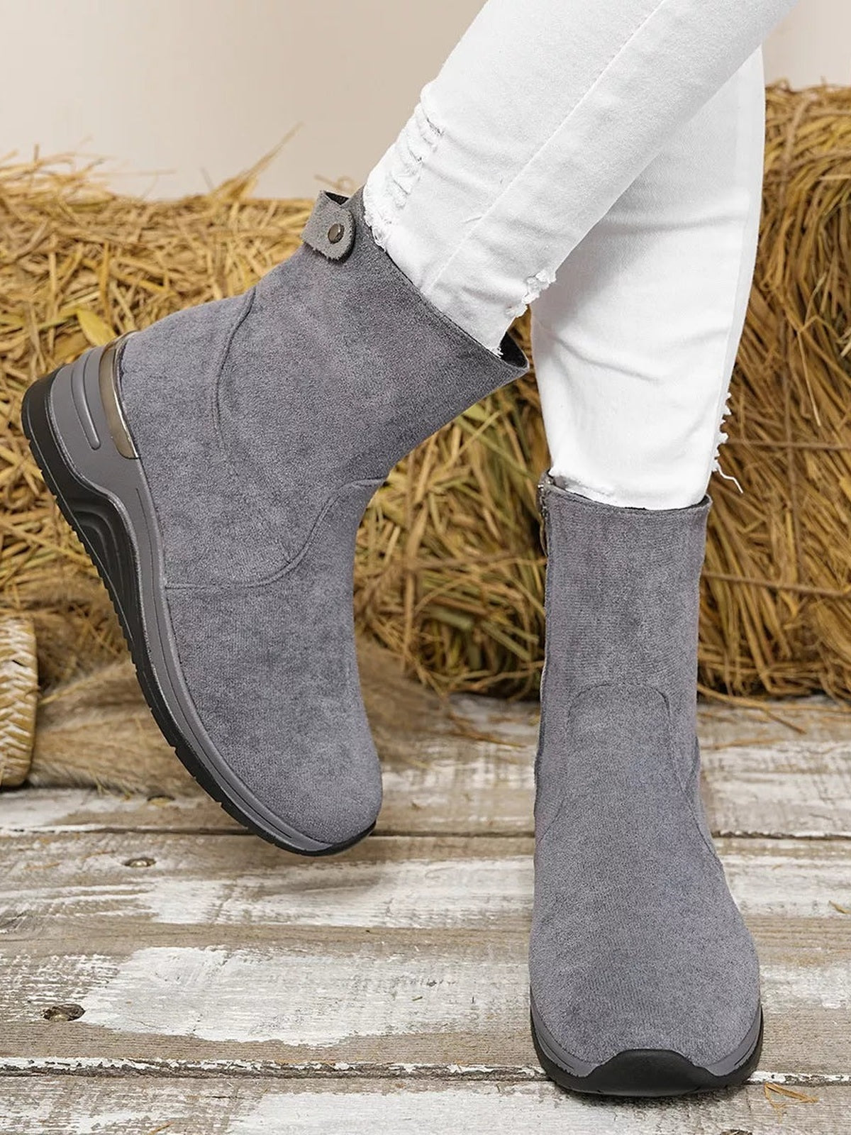 A model poses in gray suede ankle boots with rounded toes, stitched panels, black rubber soles, and mid-height wedge heels, with one foot lifted.