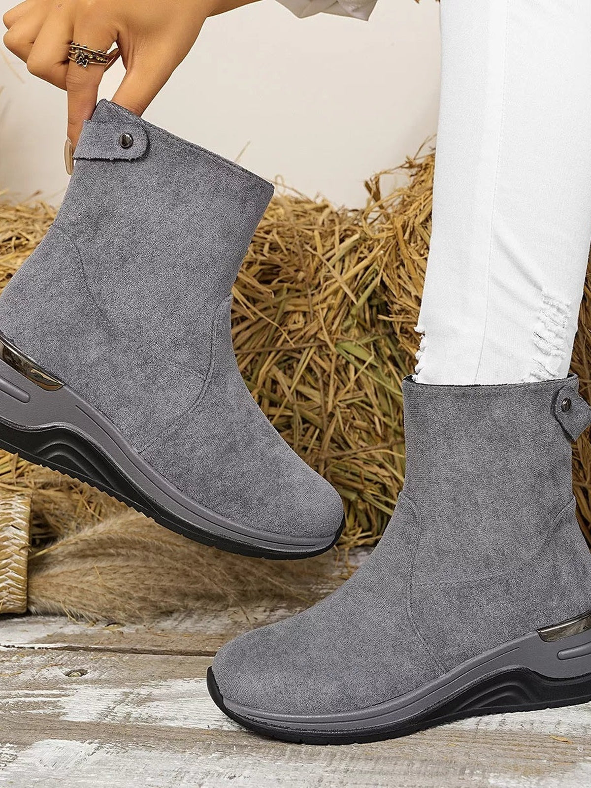 A model wears gray suede ankle boots with mid-height wedge heels, rubber soles, rounded toes, and buttoned side tabs, with one boot lifted.
