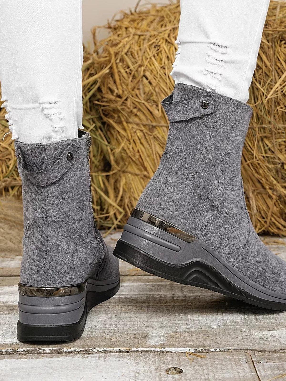 A model wears gray suede ankle boots with wraparound button tabs, metallic heel trim, raised wedge heels, and sturdy black rubber soles.