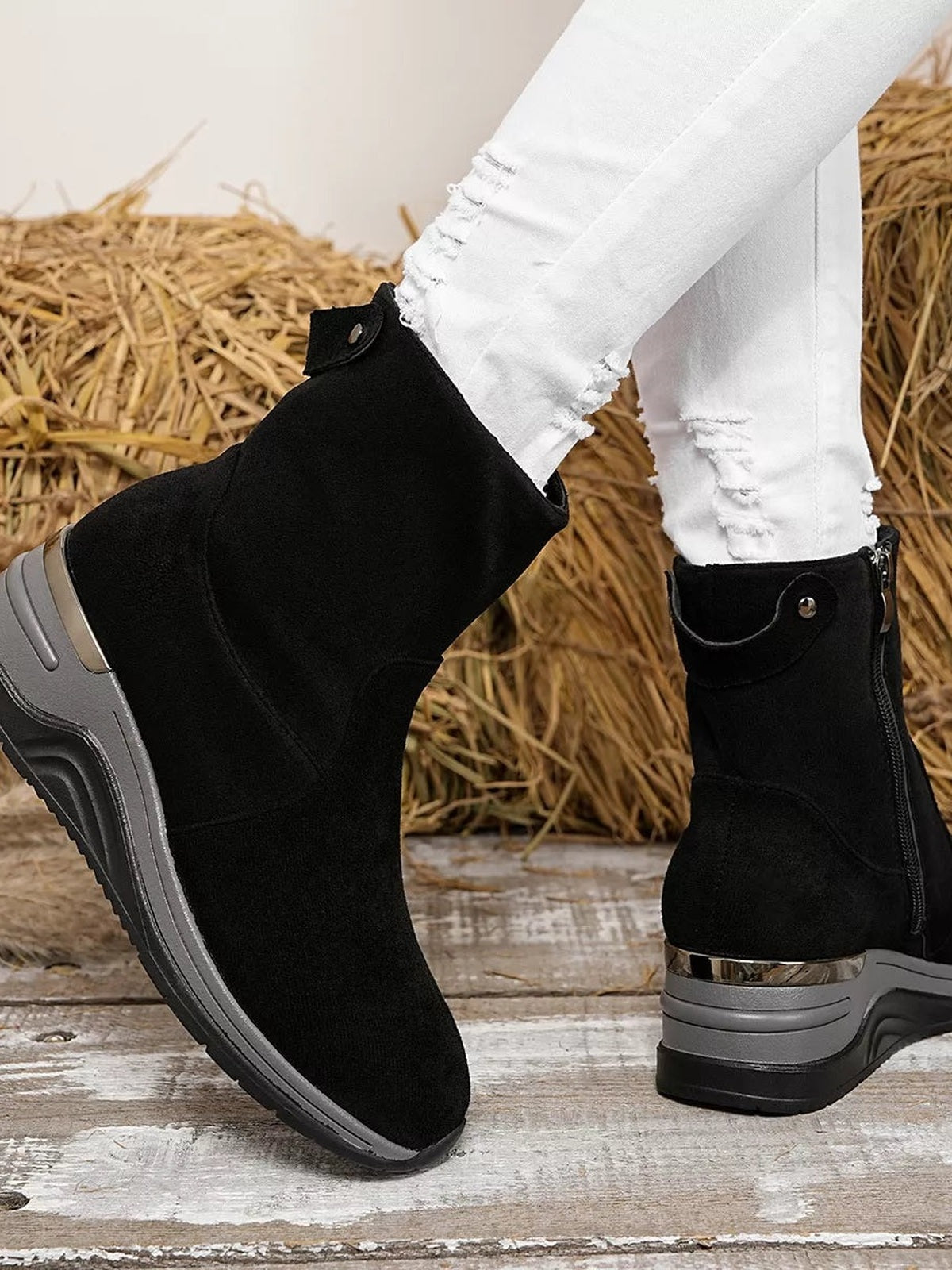 A model wears black suede ankle boots with chunky wedge heels, side zippers, metallic trim, and layered rubber soles over distressed white jeans.