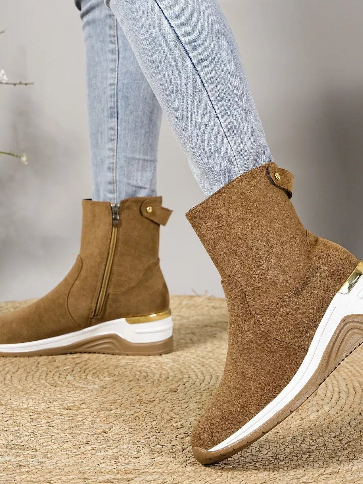 A model wears brown suede ankle boots with side zippers, buttoned ankle straps, and white rubber wedge heels, with one foot raised.