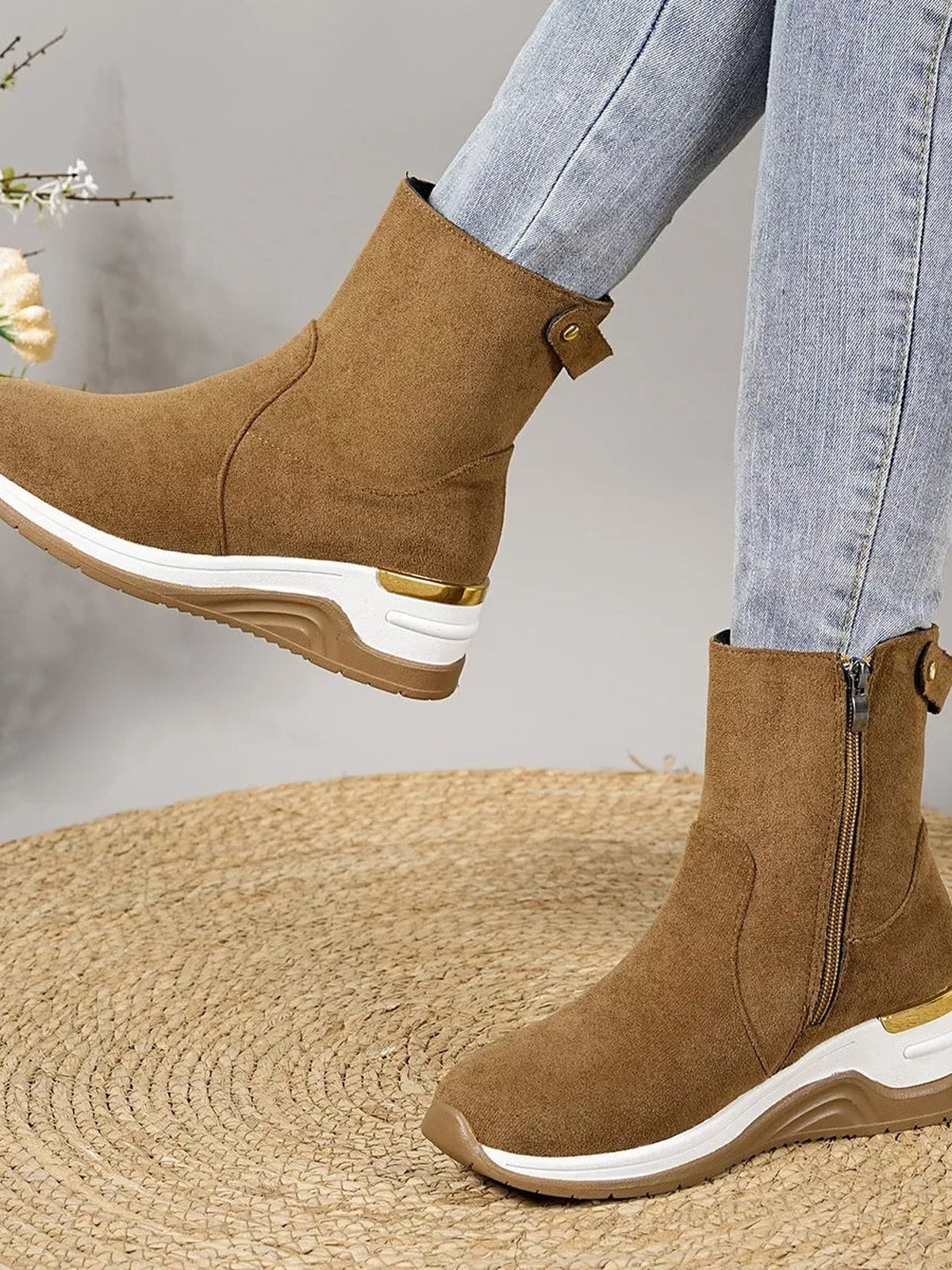 A model wears brown suede ankle boots with wedge heels, side zippers, ankle buckle straps, white midsoles, and textured rubber soles with one foot lifted.